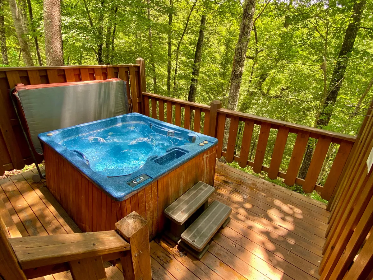 This exterior shot showcases a wooden deck nestled among the trees, with a bubbling hot tub as the focal point. The serene, natural setting is emphasized by leafy greenery and ample space for relaxation or socializing. The image suggests peaceful mornings or starlit evenings spent soaking in mountain air.