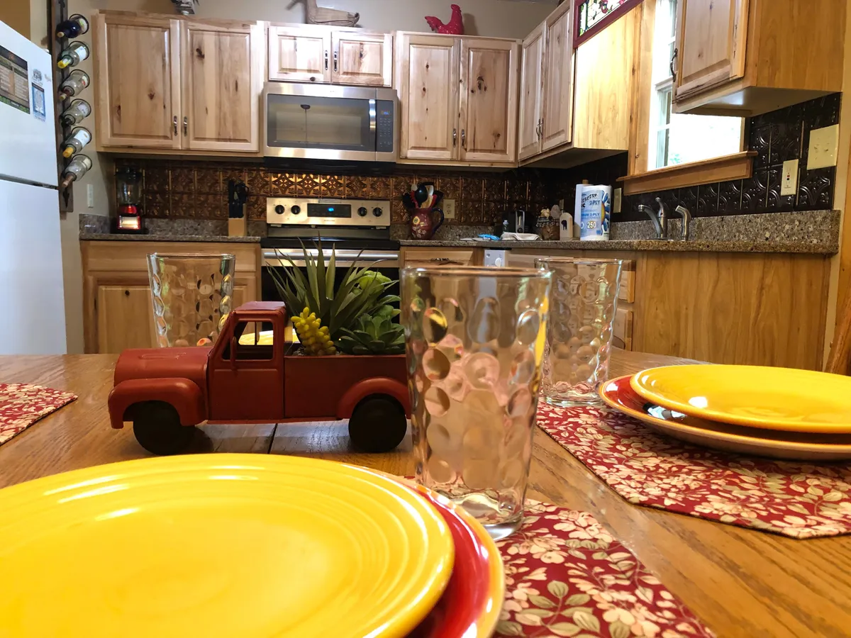 This shot focuses on the kitchen area with wooden cabinets, stainless steel appliances, colorful dinner plates, and a vintage toy truck centerpiece on the table. Glassware is neatly arranged, and the space exudes warmth and hospitality, ideal for group meals.