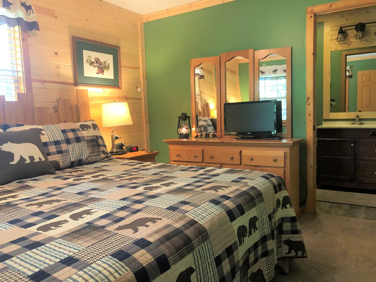 The photo features a bedroom with a patterned quilt, a wooden dresser, a mirrored vanity, a television, and framed artwork. These elements combine to showcase storage options, entertainment amenities, and thoughtful decor, all emphasizing comfort and convenience for guests.