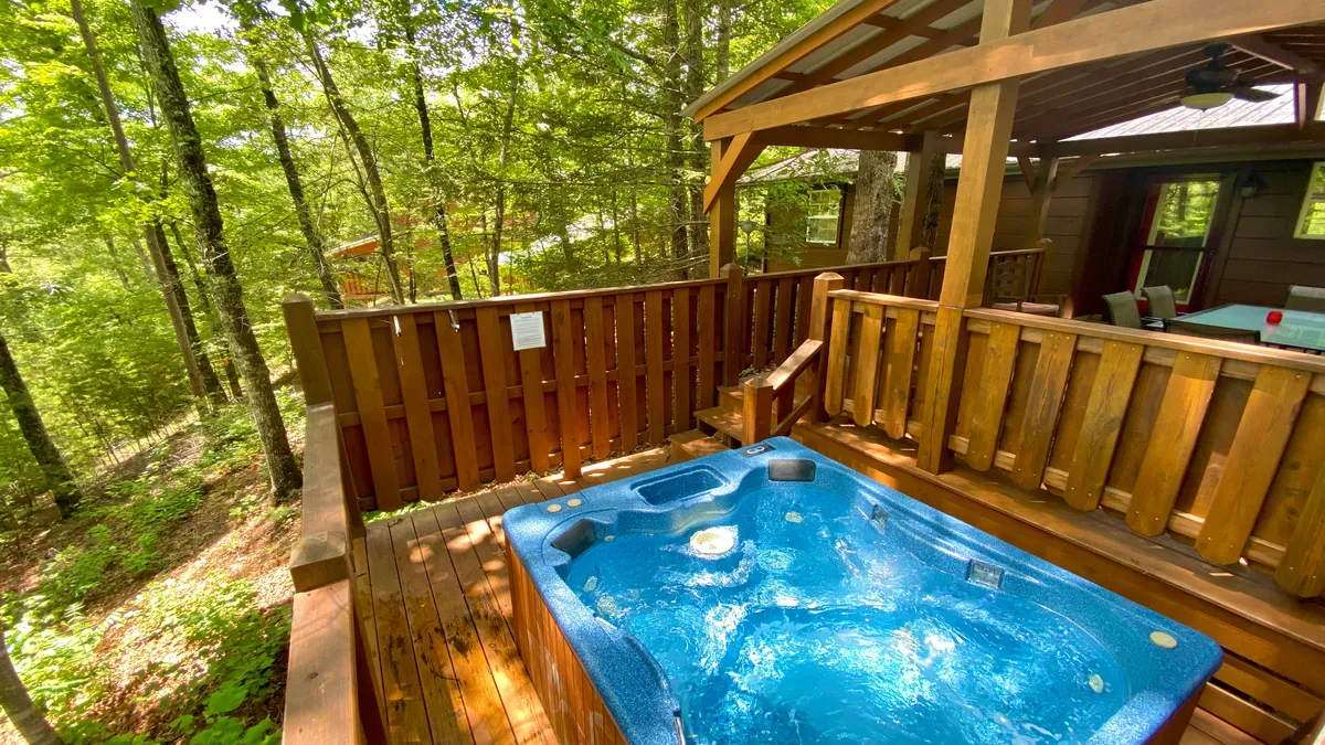 The image showcases a rustic wooden cabin exterior nestled among tall trees. A spacious deck features a bubbling hot tub as its centerpiece, with inviting seating for relaxing or entertaining. The peaceful forest surrounds create an atmosphere of privacy and serenity, perfect for unwinding in nature.