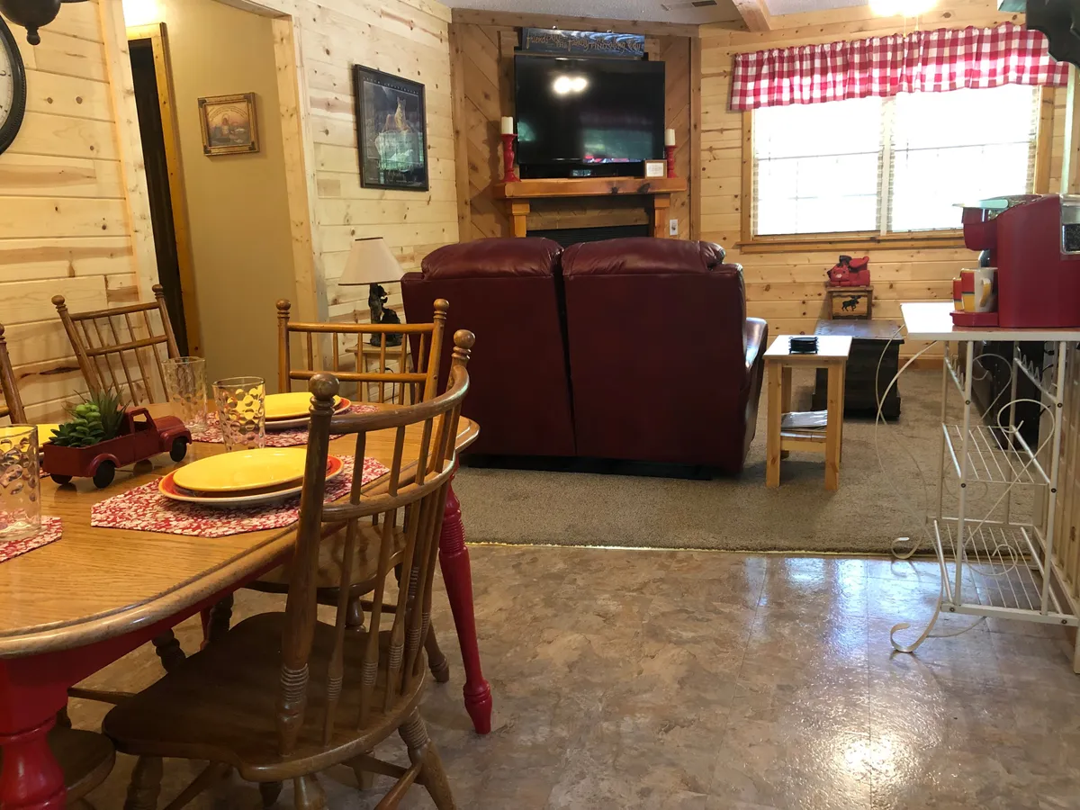 A spacious interior featuring a red leather couch, a dining table set for meals, wooden paneling, and a window with checkered curtains. The layout encourages shared meals, relaxation, and lively social moments in a welcoming atmosphere.