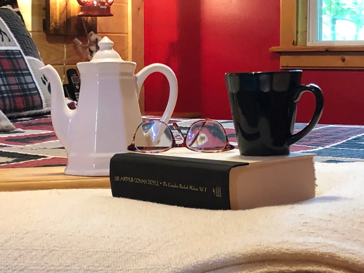 A serene moment with a white teapot, black coffee mug, and a book resting on a rustic wooden table, accented by sunglasses—implying relaxation before or after outdoor adventures.