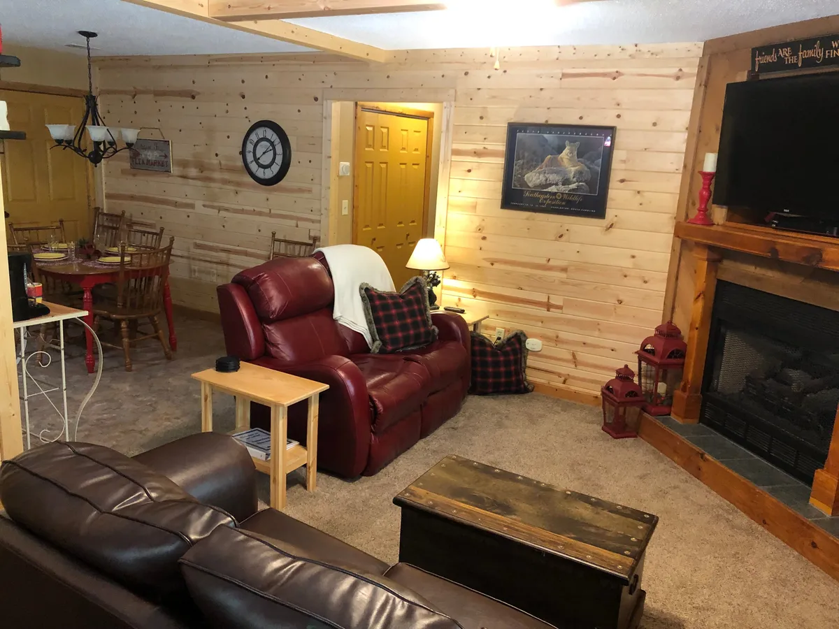A cozy living area featuring a red recliner, a rustic wooden coffee table, and a fireplace that anchors the space. The dining area is visible in the background, inviting guests to gather. Warm natural light and wood accents create a welcoming, nature-inspired atmosphere.