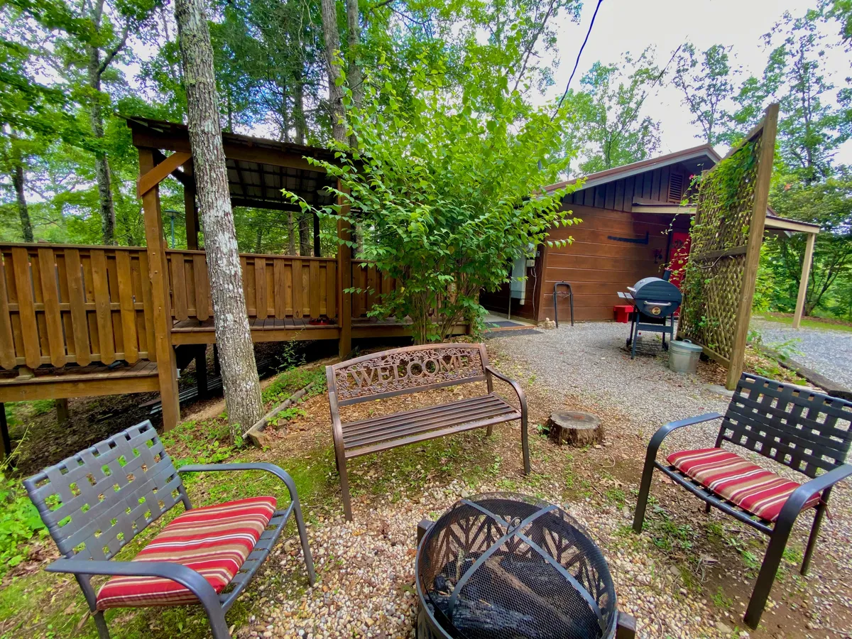 A welcoming deck scene featuring a rustic bench inscribed with 'WELCOME,' a fire pit ready for gathering, a charcoal grill, and lush greenery all around. The sunset lighting adds warmth and invites guests to imagine evenings spent sharing stories and meals.