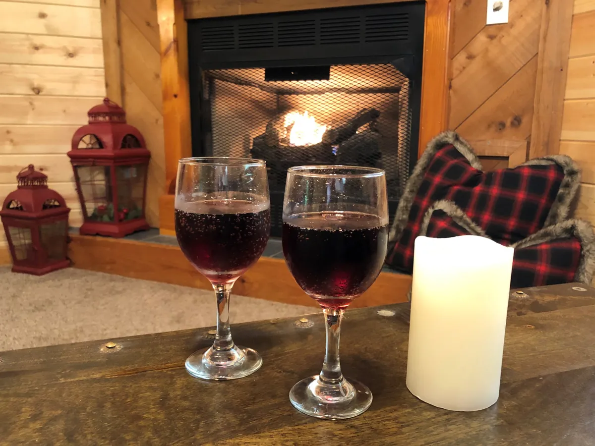 A close-up of two glasses with red drinks, a lit candle, and the fireplace glowing behind them. The scene is intimate and festive, suggesting socializing and celebrating together after exploring local attractions.