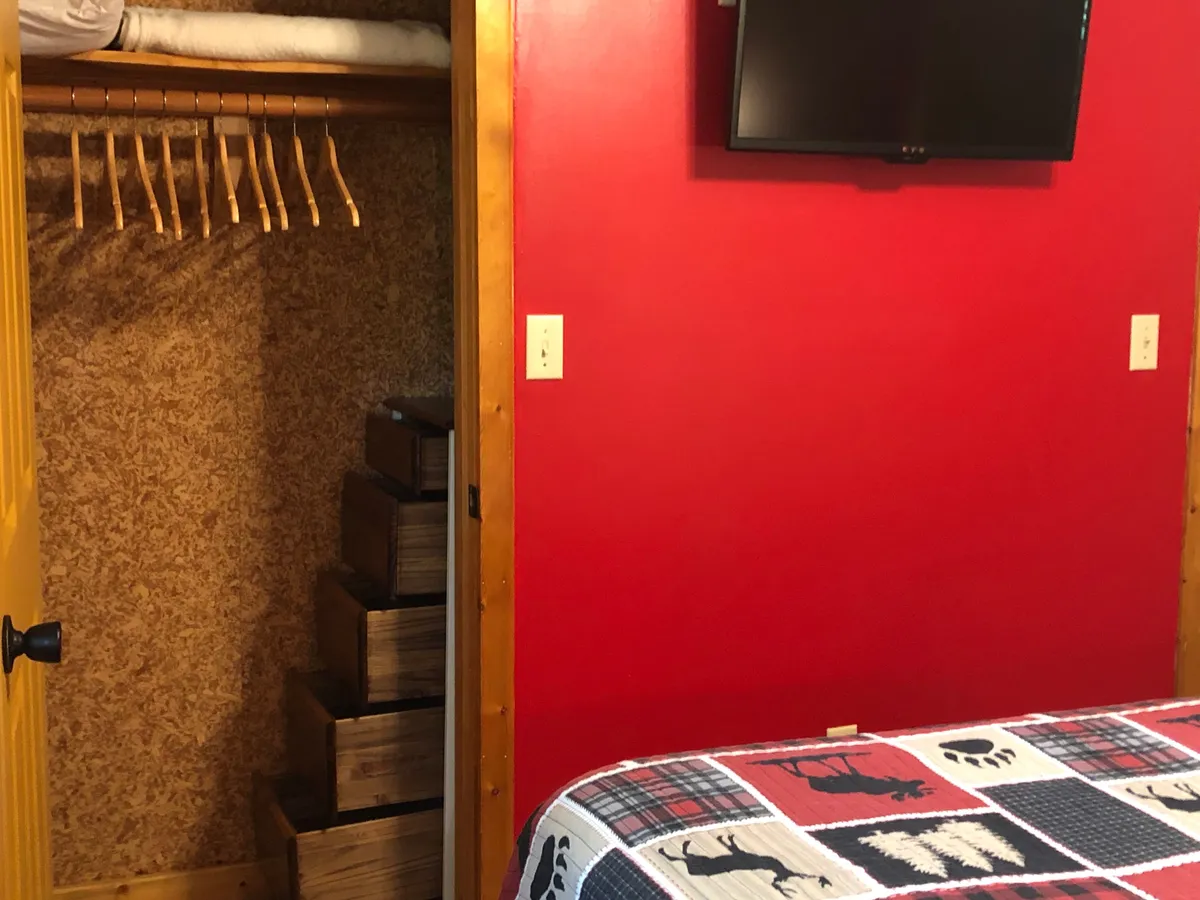 A bedroom scene highlighted by a red wall, a wooden closet, a quilted bedspread, and a TV. The setup is inviting, with clear space for unpacking and relaxing, and the presence of entertainment and storage options.