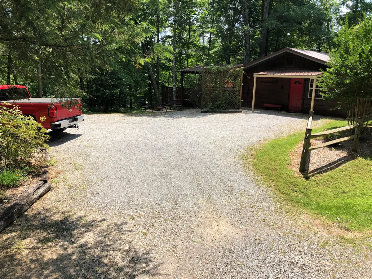 The rustic gravel driveway leads up to the cabin, with a red truck parked and a wooden structure with lattice visible. Leafy green foliage frames the scene, emphasizing the cabin’s mountain setting and easy access for exploration.