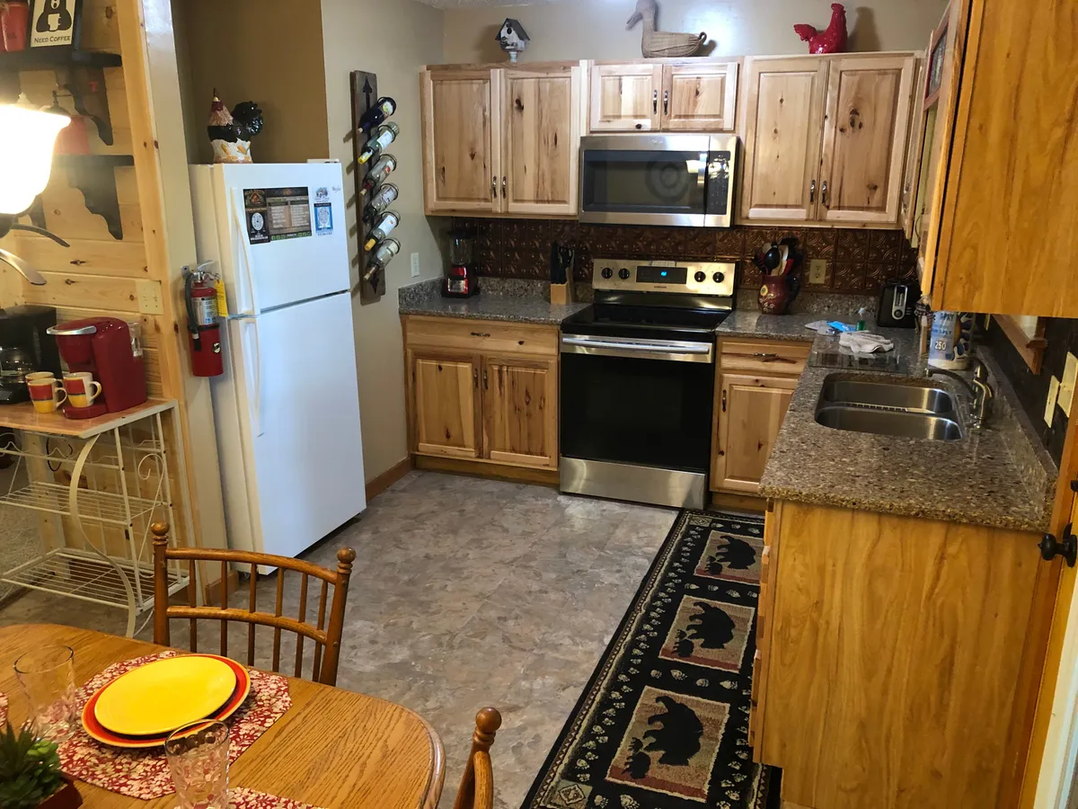 This image showcases a warm and welcoming kitchen and dining area. Visible are wooden cabinets, stainless steel appliances, a spacious dining table, and a bear-themed rug, all set in an open-concept layout. The space appears ready for cooking, family gatherings, and shared meals.