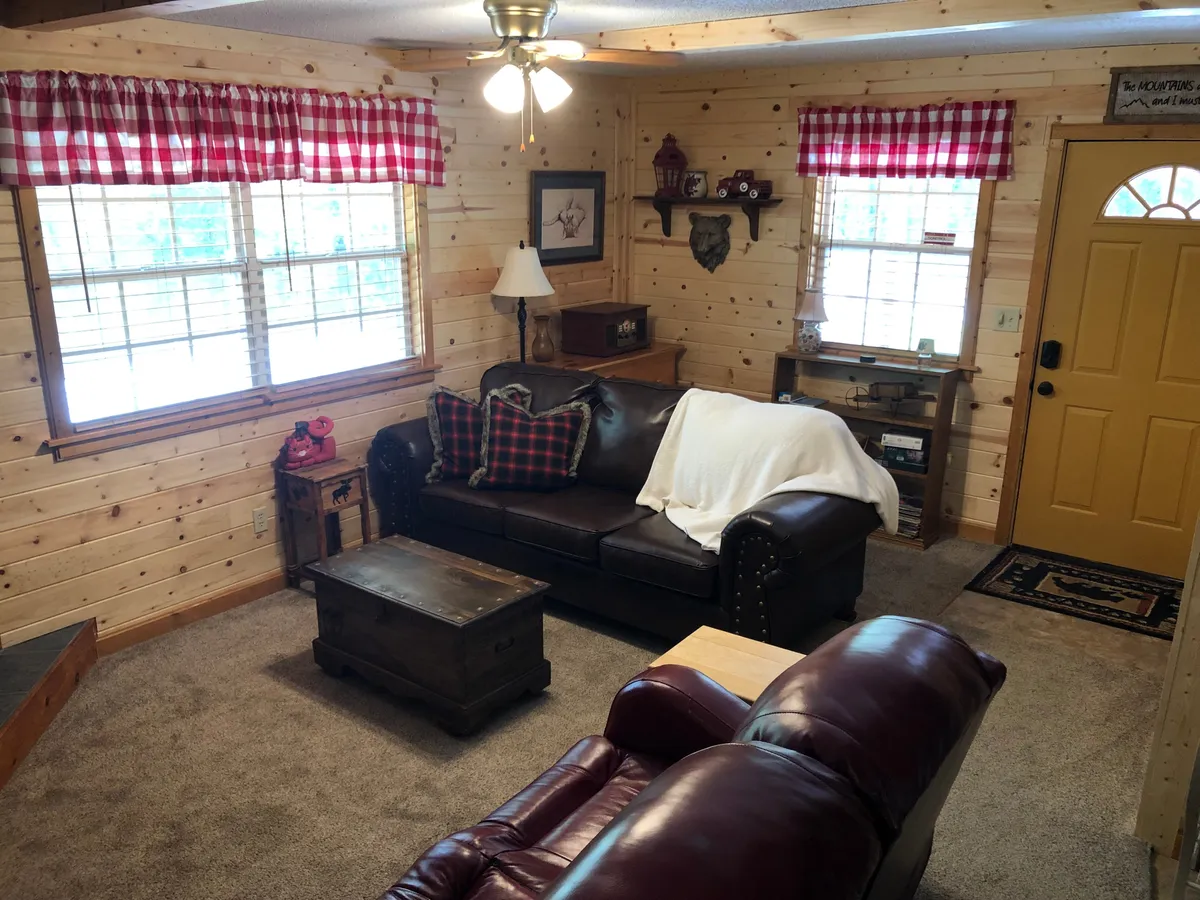 A warm, inviting living room featuring a brown leather sofa with plaid pillows, a cozy wooden coffee table, and a bright yellow front door, all bathed in natural light from windows with checkered curtains. The space exudes rustic charm and a sense of welcome, perfect for relaxing or socializing upon arrival.